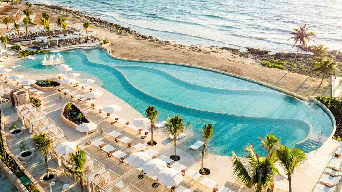 En stor pool omgivet af solsenge og parasoller ligger ved stranden med udsigt over havet. (AI-genereret tekst)