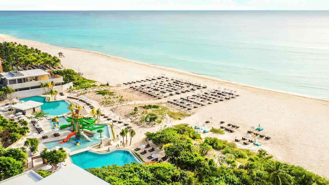 Billede af en sandstrand ved havet med blåt vand. På stranden er der liggestole og parasoller. I forgrunden ses en pool med vandrutsjebaner samt omgivelser med grønne planter. (AI-genereret tekst)