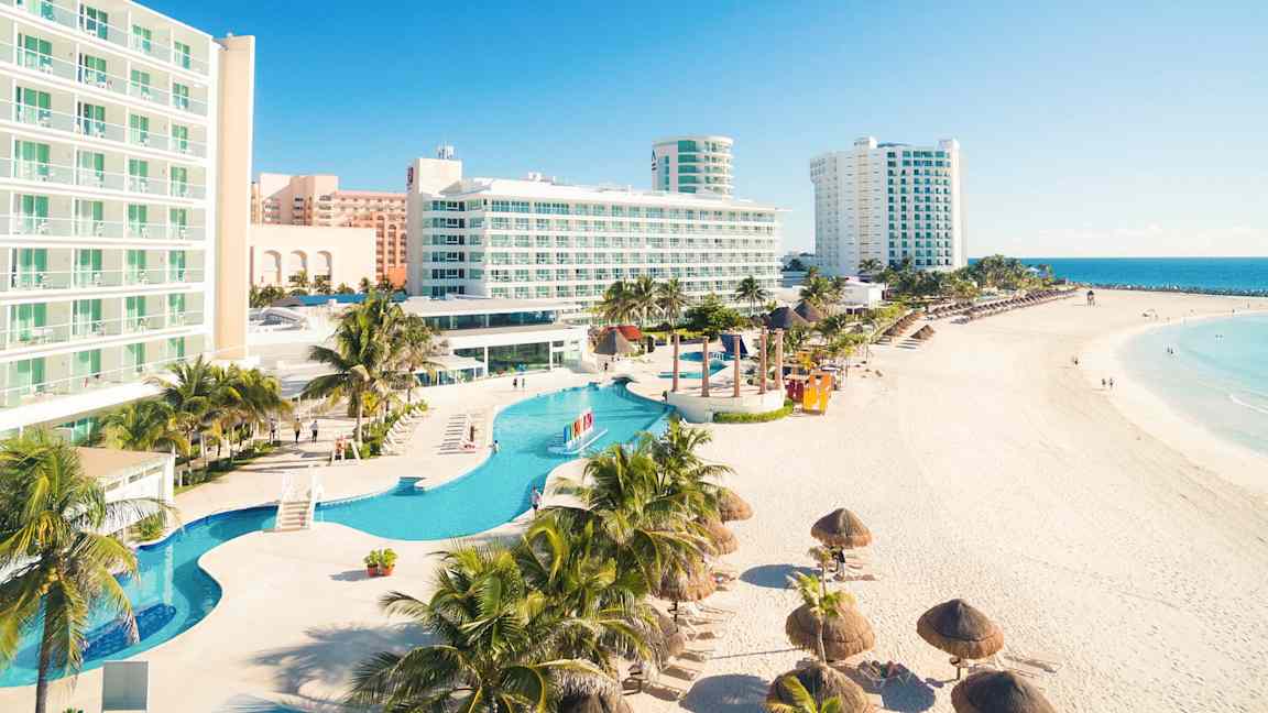 Et hotelkompleks ved en strand med pools i forgrunden og havudsigt i baggrunden. (AI-genereret tekst)