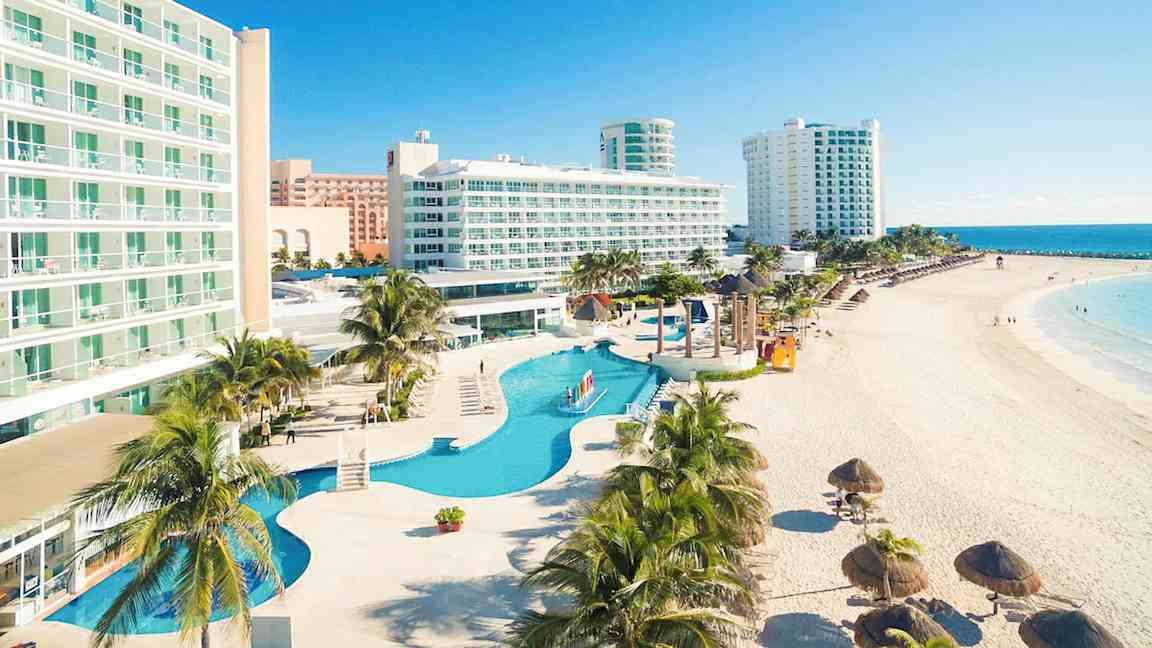 Billede af hotel ved stranden med pool og palmer. (AI-genereret tekst)