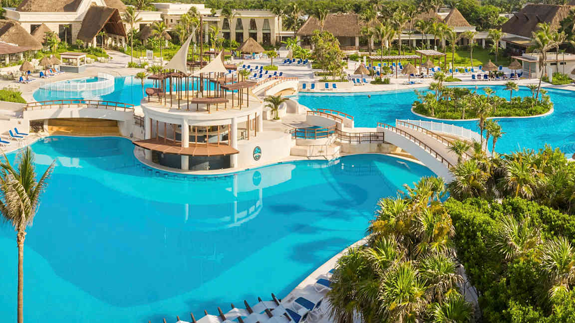 En stor pool med broer og et centralt område omgivet af tropiske planter og bygninger. (AI-genereret tekst)