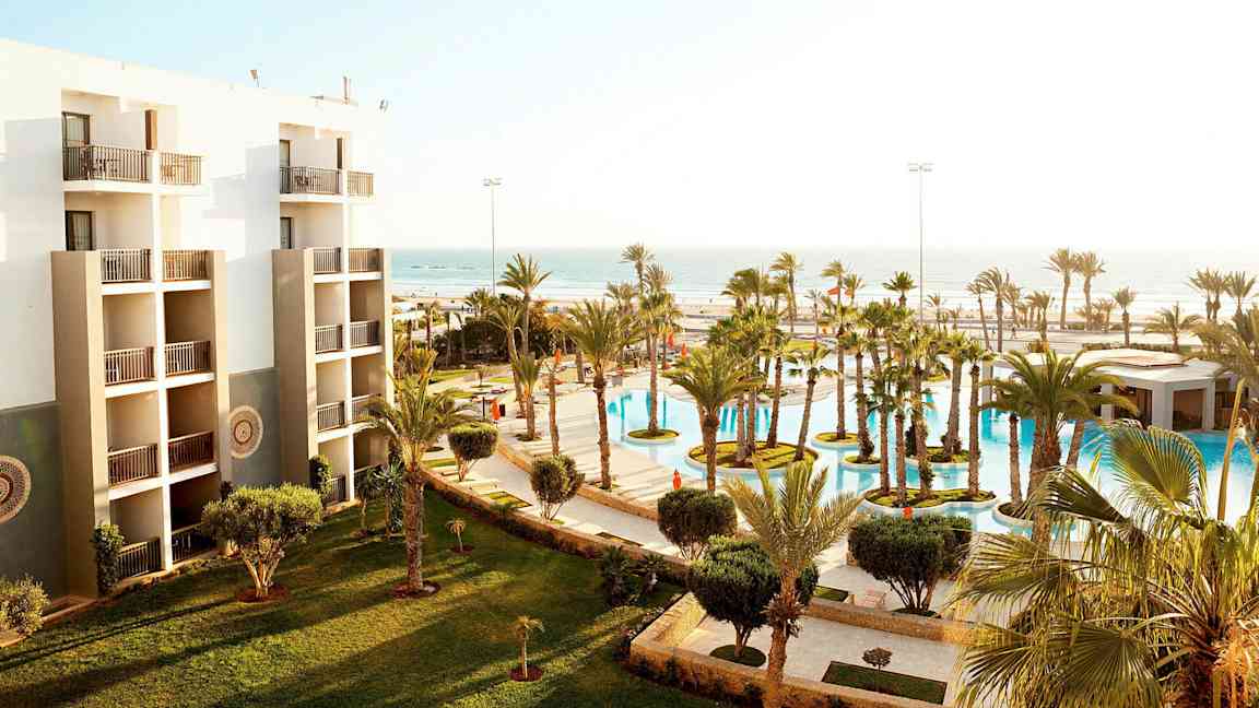 Hotellets facade med altaner og en udsigt over frodige haver og en pool samt havet i baggrunden. (AI-genereret tekst)