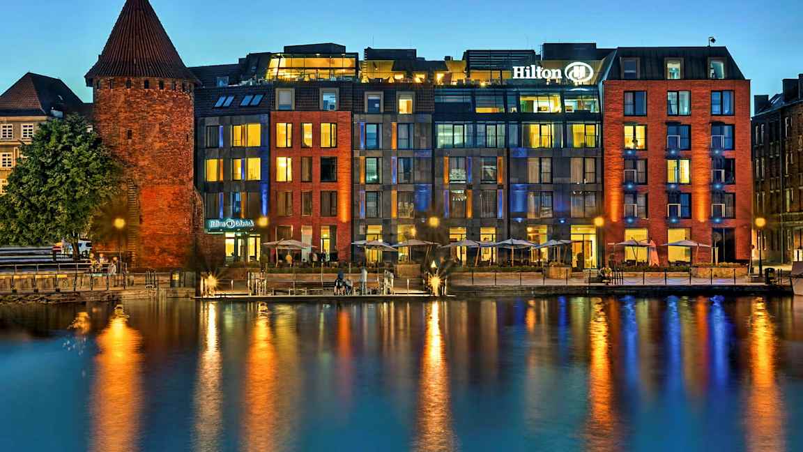 En historisk murbygning ved siden af et moderne hotel ved vandet om aftenen. (AI-genereret tekst)