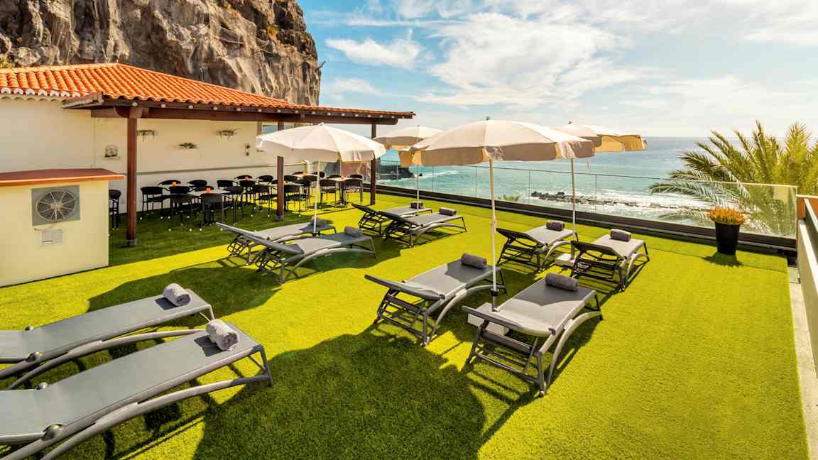 En terrasse med liggestole og parasoller på en græsplæne, ved et lavt hus med et orange tegltag. Udsigt over havet og en klippevæg i baggrunden. (AI-genereret tekst)