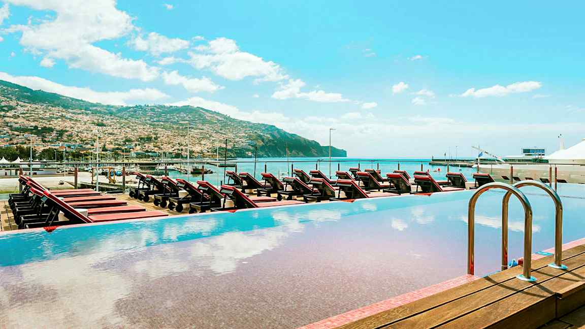 Infinitypool med udsigt over liggestole, havet og det omkringliggende bjerglandskab. (AI-genereret tekst)