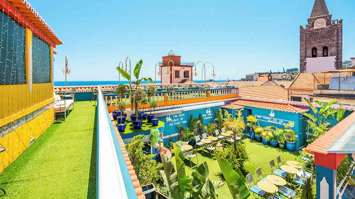 En tagterrasse med grønne planter, siddepladser og farverige bygninger i baggrunden. (AI-genereret tekst)