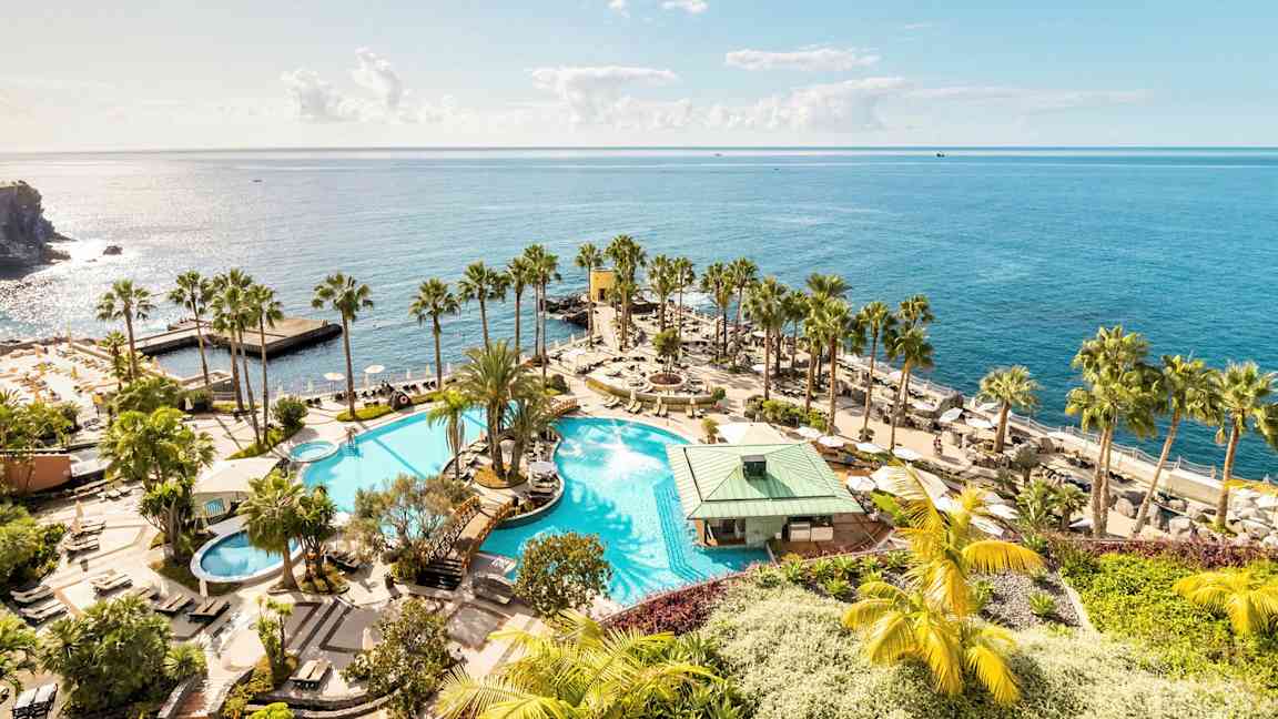 En stor pool med omkringliggende palmer og solsenge, beliggende ved stranden med udsigt over det glitrende hav. (AI-genereret tekst)