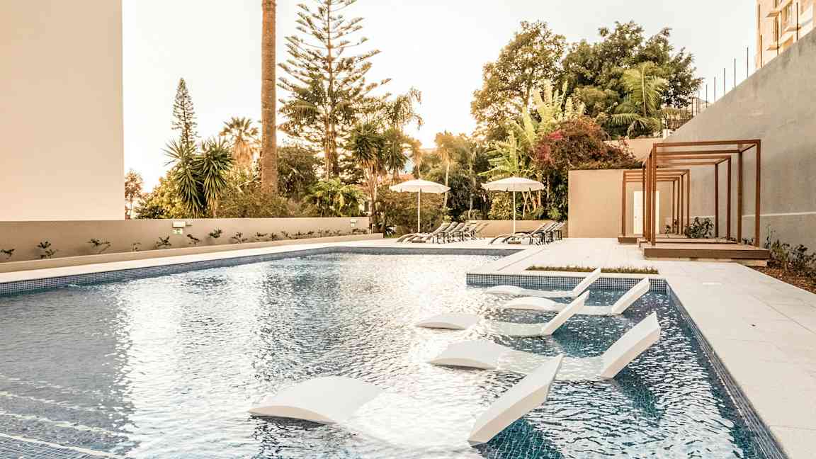 Udendørs pool med solsenge og parasoller, omgivet af træer og vegetation. (AI-genereret tekst)