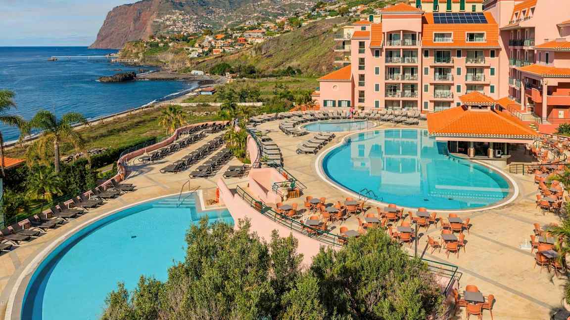 Et hotelområde ved kysten med to store pools, liggestole, borde og omkringliggende bygninger med orange tage. Baggrunden viser en klippe ved havet og boligområder langs skråningen. (AI-genereret tekst)