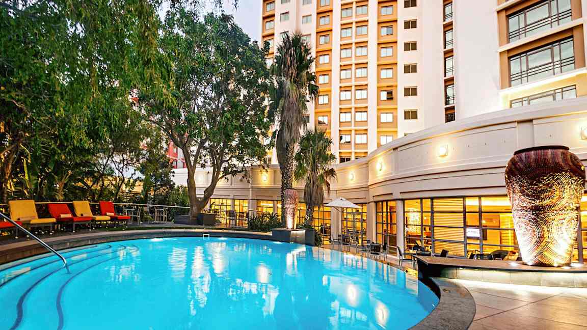 En oplyst udendørs pool omgivet af grønne planter og en bygning i baggrunden. (AI-genereret tekst)