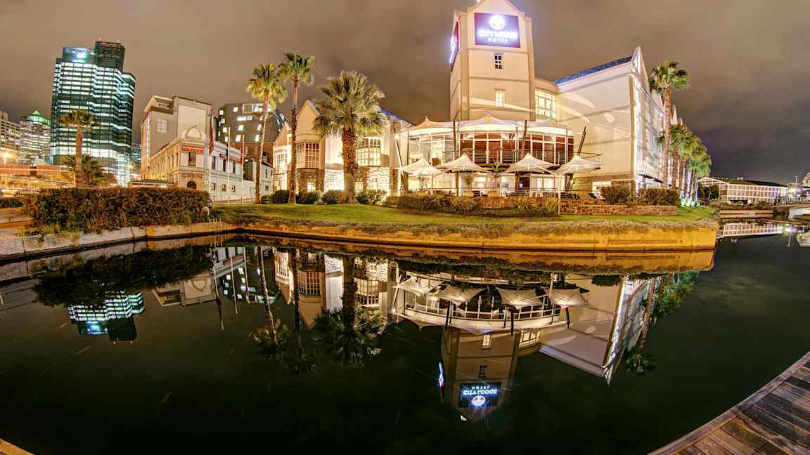 Hotel og bygninger ved en kanal om aftenen med spejlreflekser i vandet. (AI-genereret tekst)