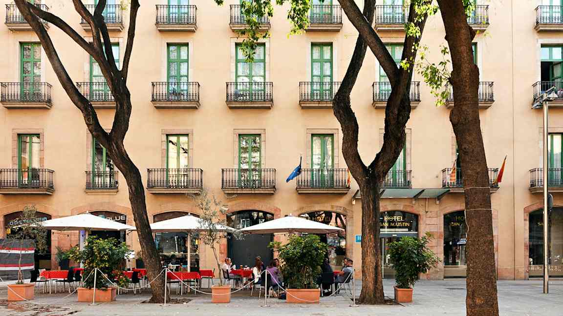 Et hotel med flere etager og altaner, et fortov med parasoller og siddepladser mellem træer. (AI-genereret tekst)