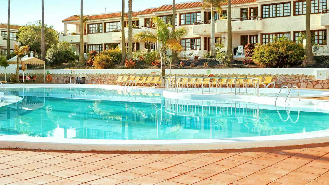 En swimmingpool omgivet af liggestole med en hvid bygning i baggrunden. (AI-genereret tekst)