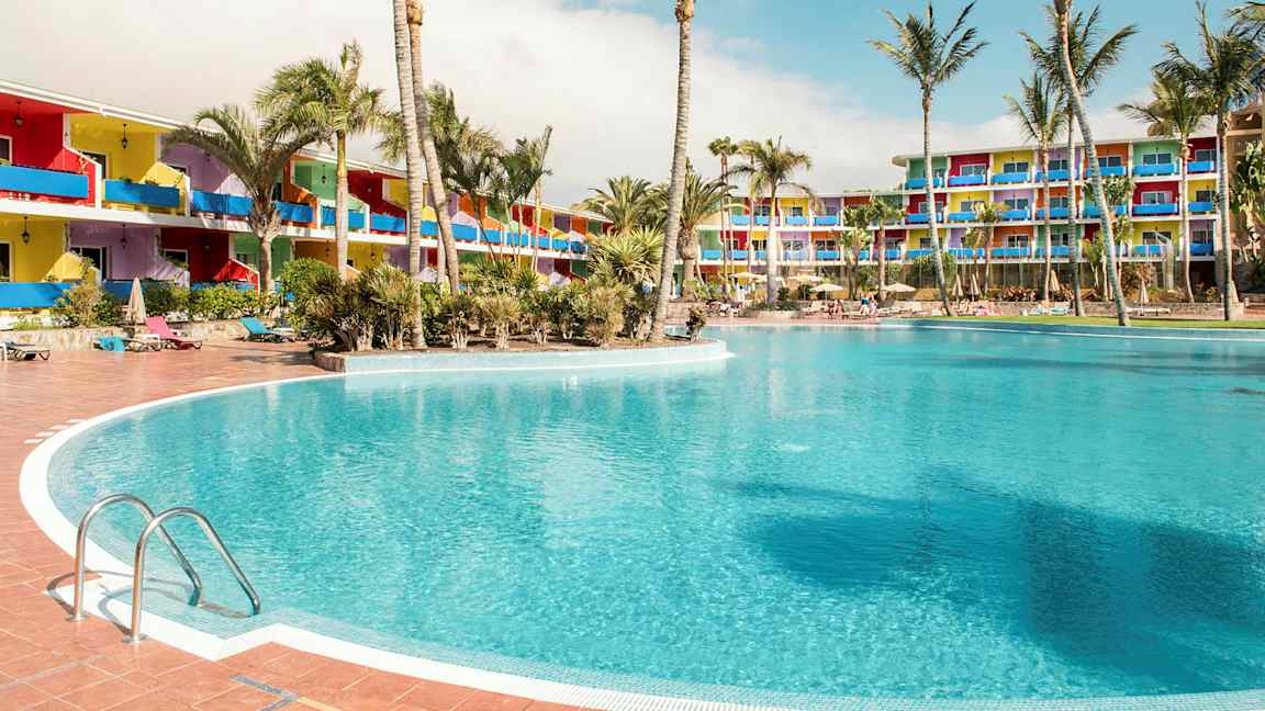 En stor pool omgivet af palmer, med farverige hotelbygninger i baggrunden. (AI-genereret tekst)
