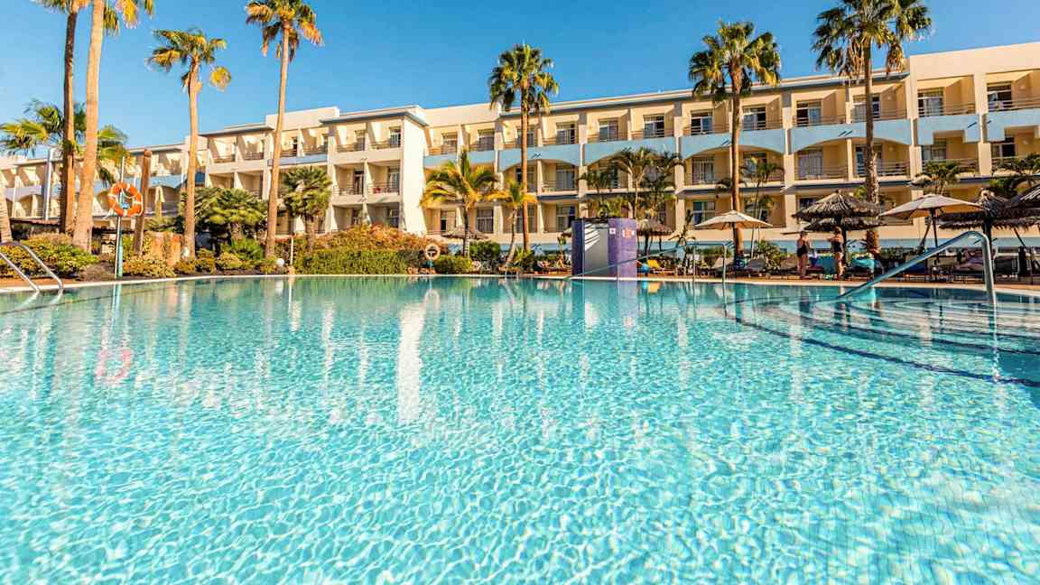 En udendørs pool omgivet af palmer og et hotel i baggrunden. (AI-genereret tekst)