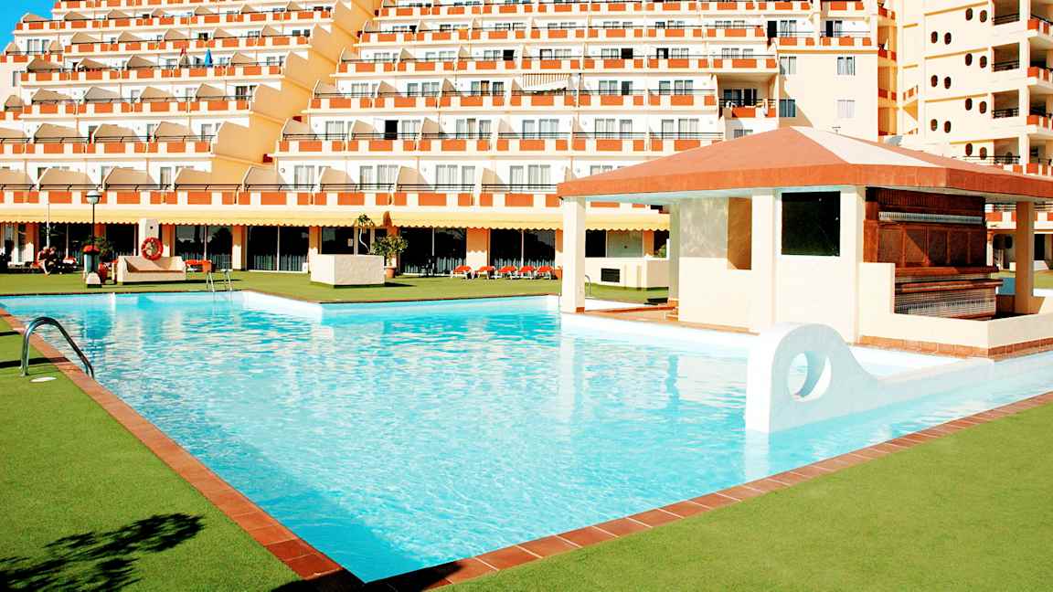 En stor pool omgivet af grønne områder og et hotel med flere etager i baggrunden. (AI-genereret tekst)