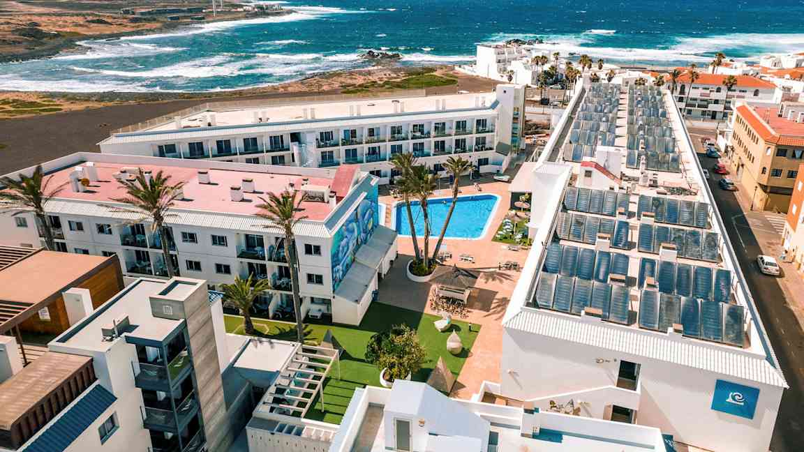 En hotelresort med pool, omgivet af bygninger, med udsigt til havet og bølgerne. (AI-genereret tekst)