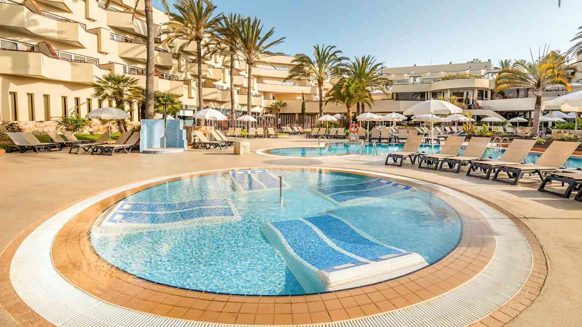 Et poolområde på et hotel med liggestole og parasoller omkring poolen, omgivet af palmer og altaner. (AI-genereret tekst)