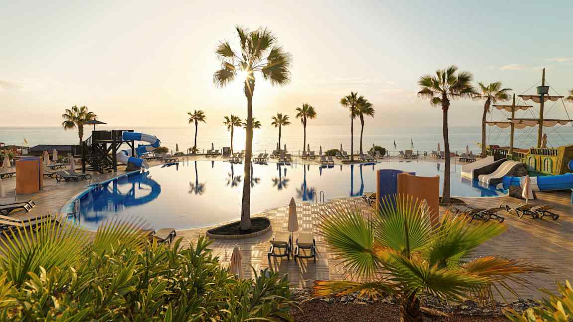 Billede af en pool med vandrutsjebaner, solsenge og palmer samt havet i baggrunden ved solnedgang. (AI-genereret tekst)