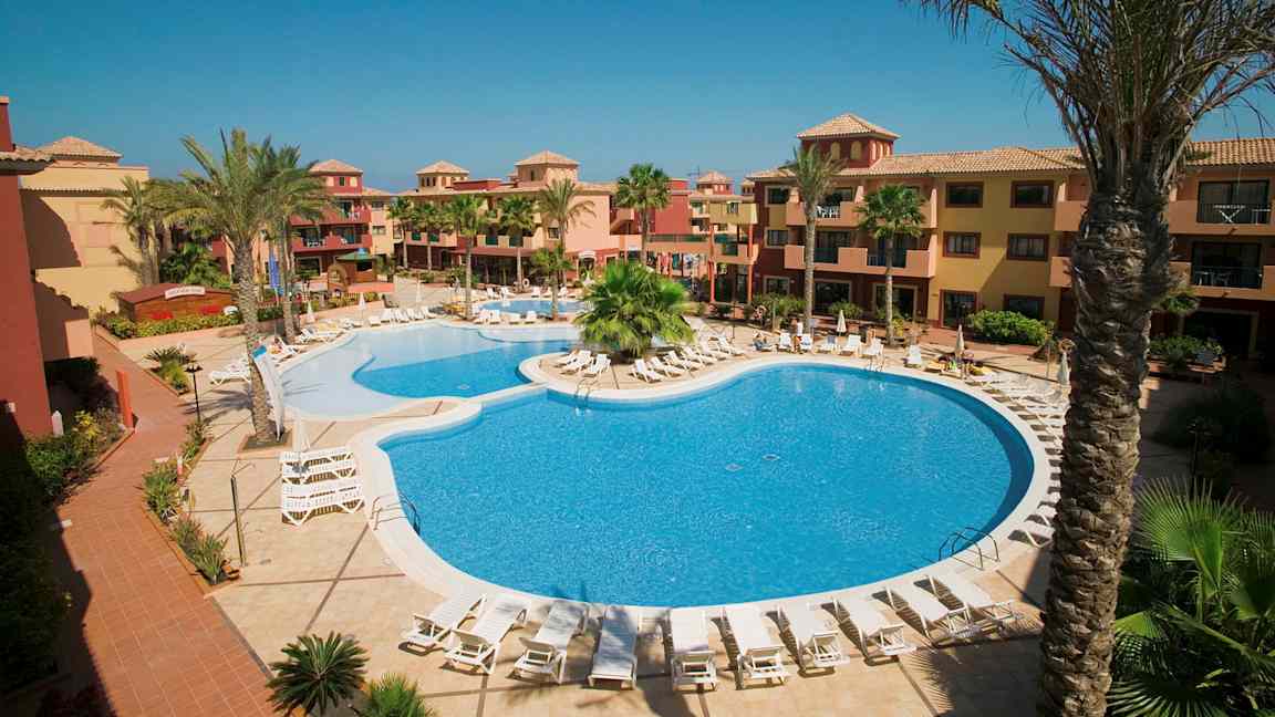 Hotelområde med pool og liggestole, omgivet af palmer og bygninger i varme farver. (AI-genereret tekst)