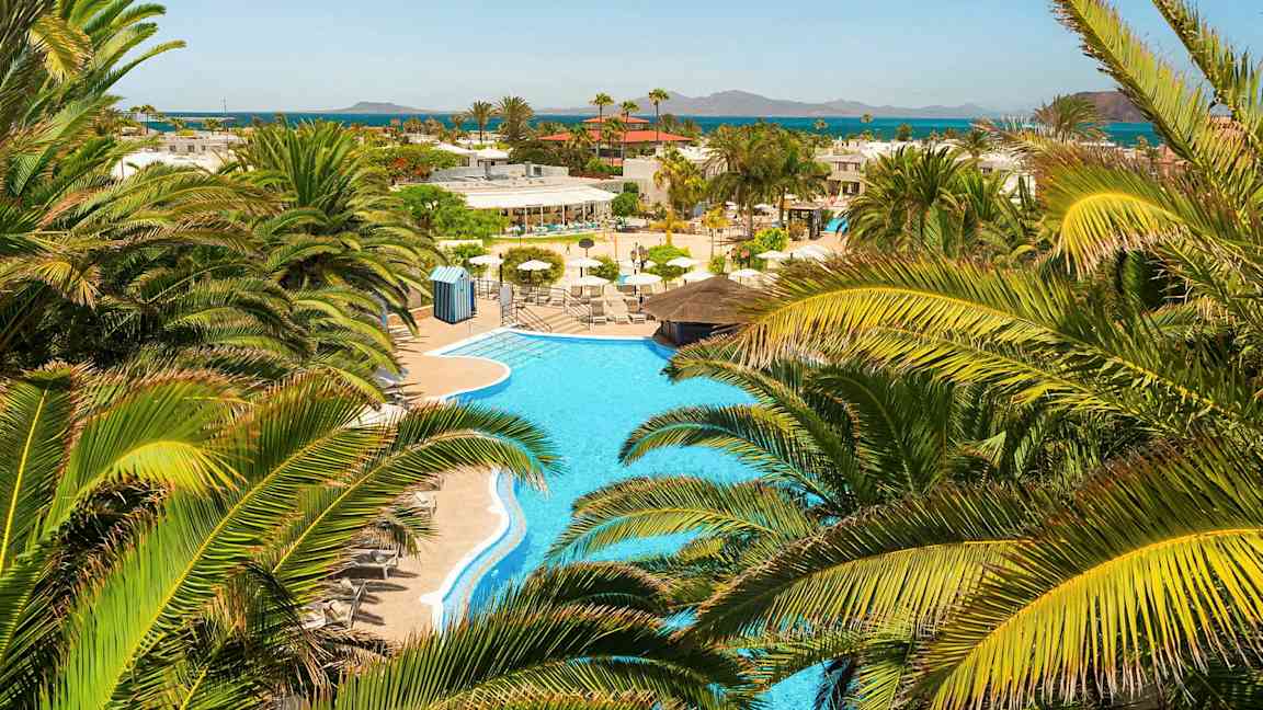 Et udsyn over en swimmingpool omgivet af palmer med udsigt til havet i det fjerne. (AI-genereret tekst)