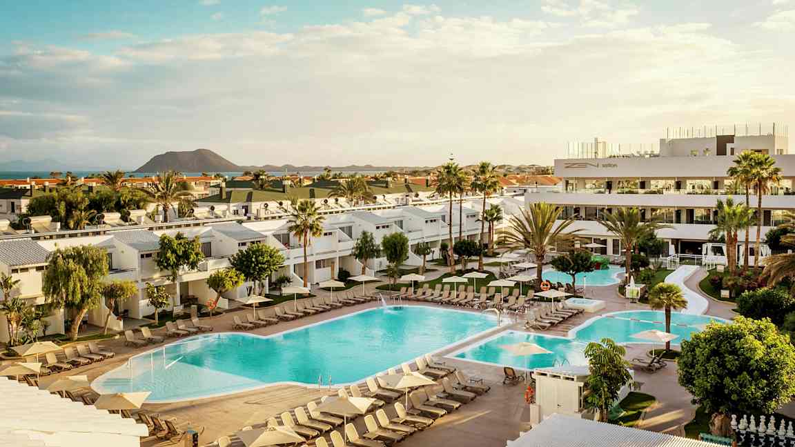 Billede af et hotelområde med pools, liggestole og omkringliggende bygninger samt en vulkan i baggrunden. (AI-genereret tekst)