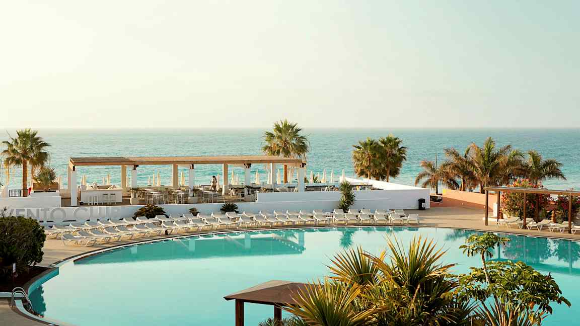 Hotelområde med swimmingpool, liggestole, palmer og havudsigt. (AI-genereret tekst)
