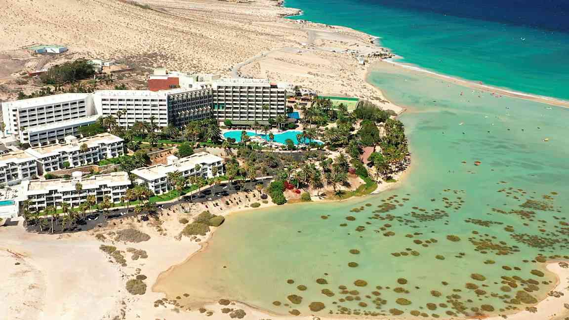 Hotelkompleks ved en strand med turkis vand og omgivelser af ørkenlandskab. (AI-genereret tekst)