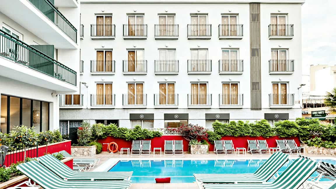 Billede af en hotelbygning med hvid facade og flere altaner. Foran bygningen er en udendørs pool med stribede liggestole placeret omkring. (AI-genereret tekst)