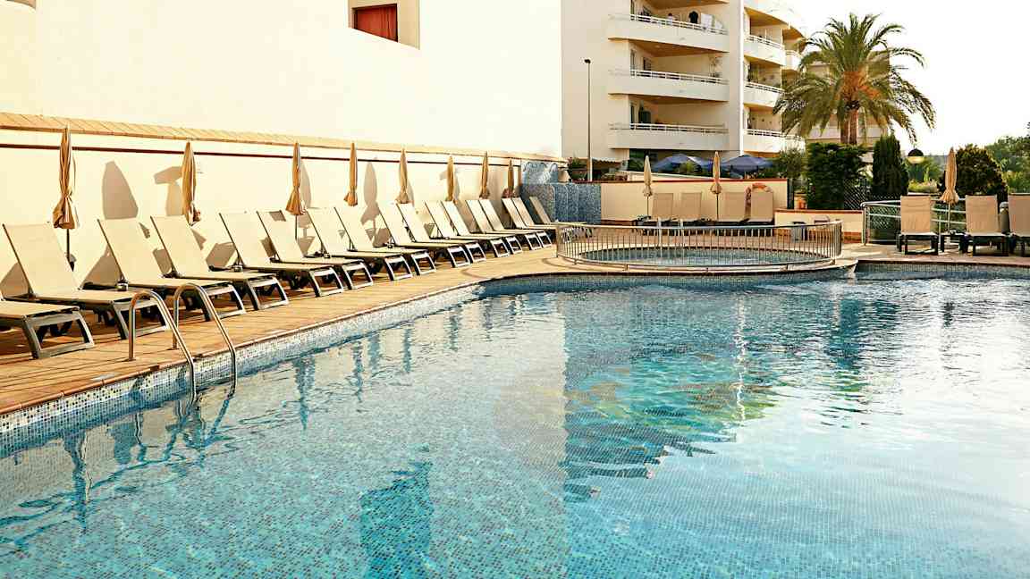 En udendørs swimmingpool omgivet af liggestole og parasoller, med en bygning og en palme i baggrunden. (AI-genereret tekst)