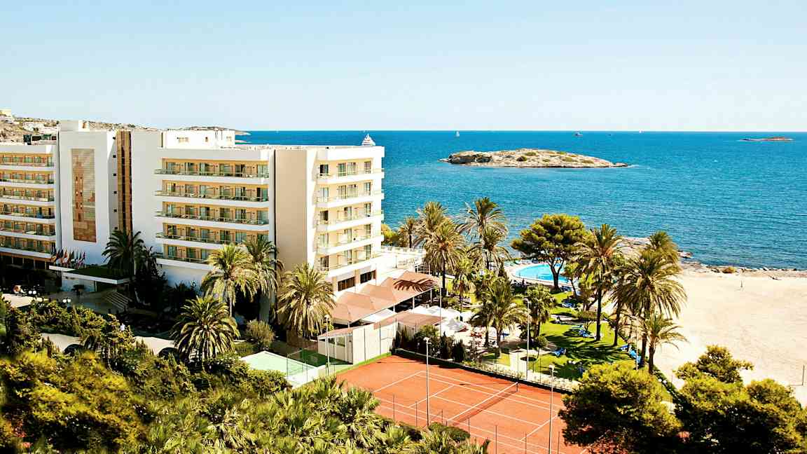 Billede af et feriested ved stranden med en bygning, palmer, swimmingpool og tennisbaner, placeret ved havet. (AI-genereret tekst)