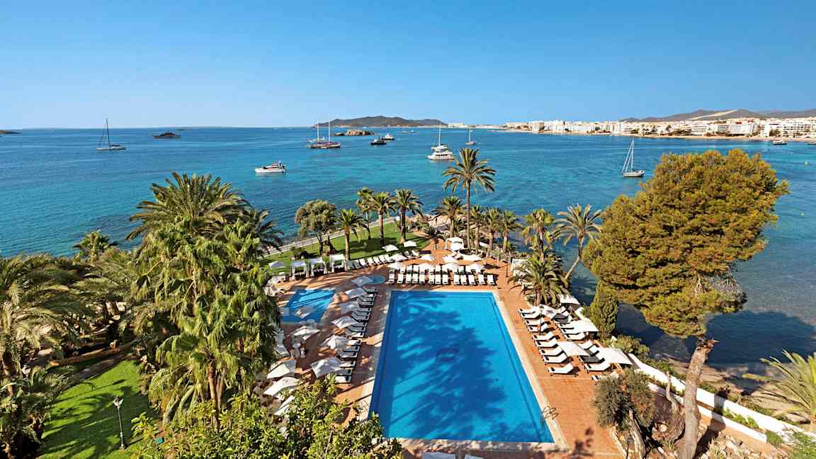 En pool og solsenge omgivet af palmer ved kystlinjen med en baggrund af et blåt hav og sejlbåde. (AI-genereret tekst)