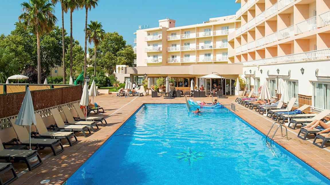 En hotelbygning ses ved siden af en udendørs pool med liggestole og parasoller omkring. (AI-genereret tekst)