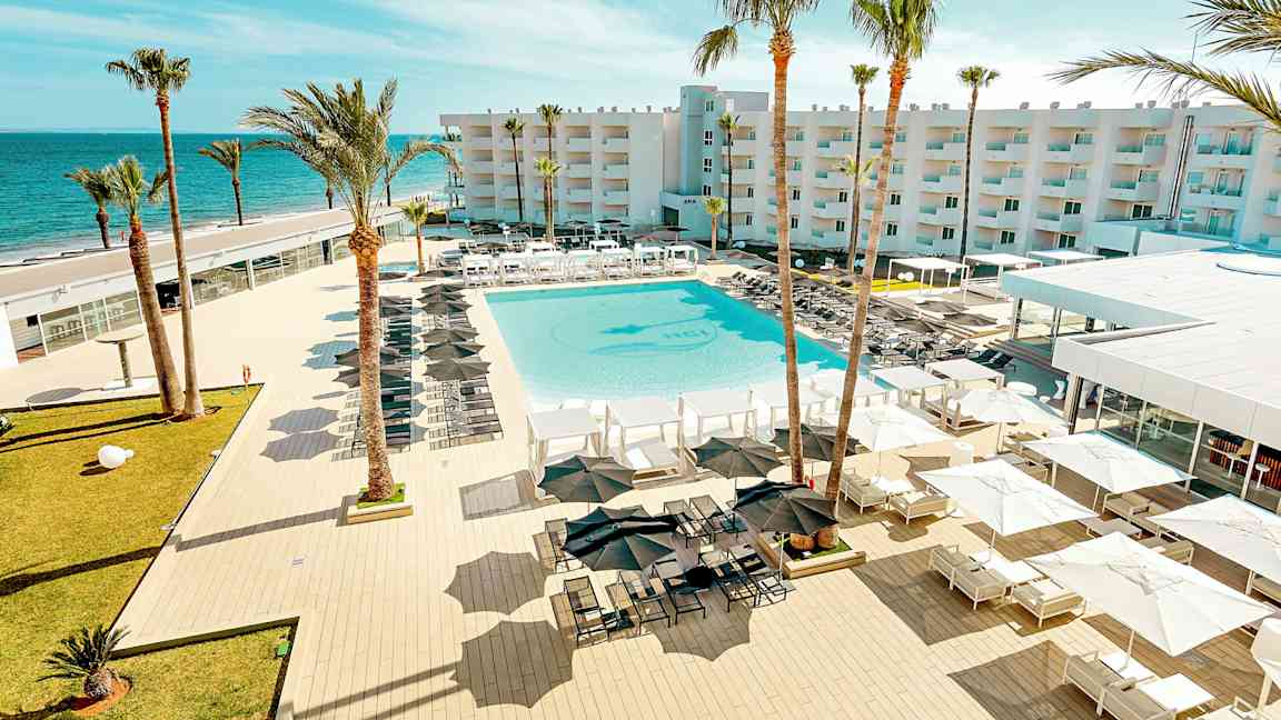 En udsigt over et feriested med en stor pool, omgivet af liggestole og parasoller, med palmer og en bygning i baggrunden tæt på havet. (AI-genereret tekst)