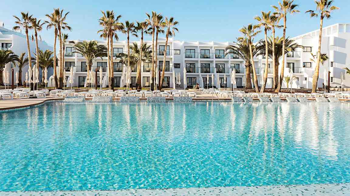 Hotellets poolområde med klart blåt vand, omgivet af liggestole, parasoller og palmer. I baggrunden ses en hvid bygning med altaner. (AI-genereret tekst)