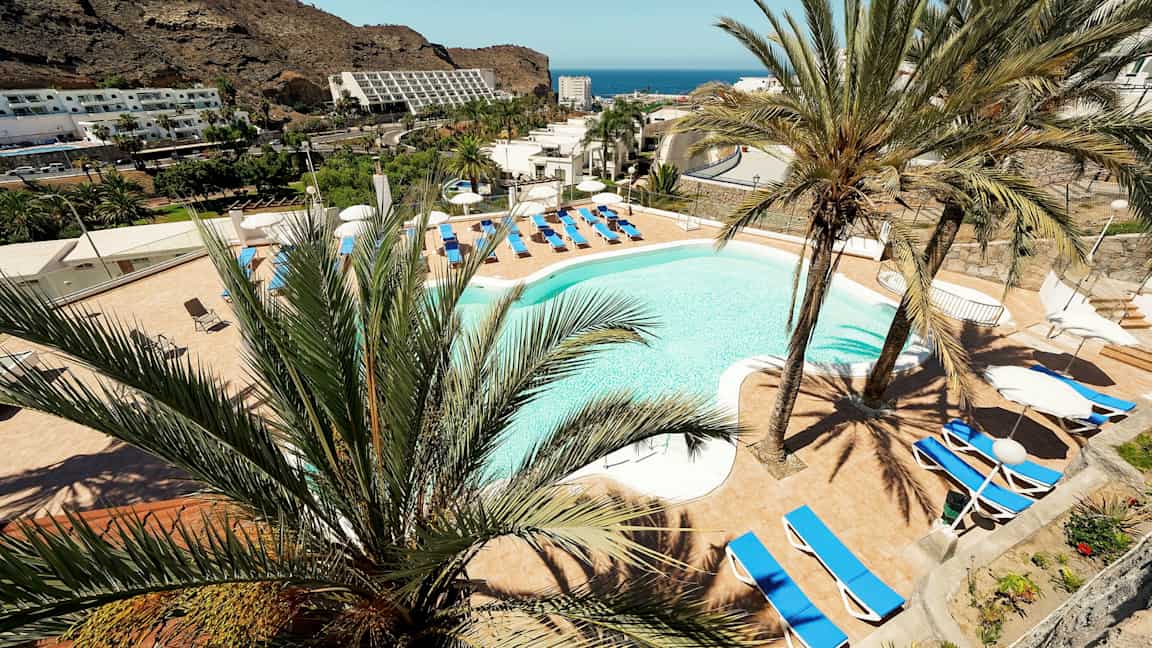 Udsigt over en hotelpool omgivet af liggestole og palmer med bjerge og hav i baggrunden. (AI-genereret tekst)