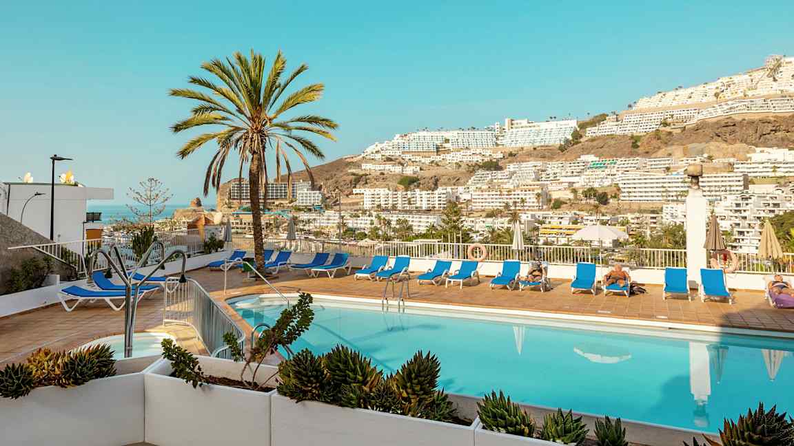 Udendørspool med liggestole og palmer samt udsigt over bjerge og bygninger. (AI-genereret tekst)