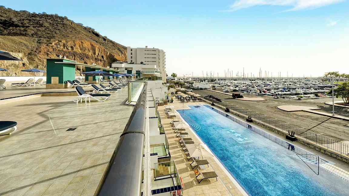 Poolområde med liggestole ved siden af en marina og en bjergskråning. (AI-genereret tekst)