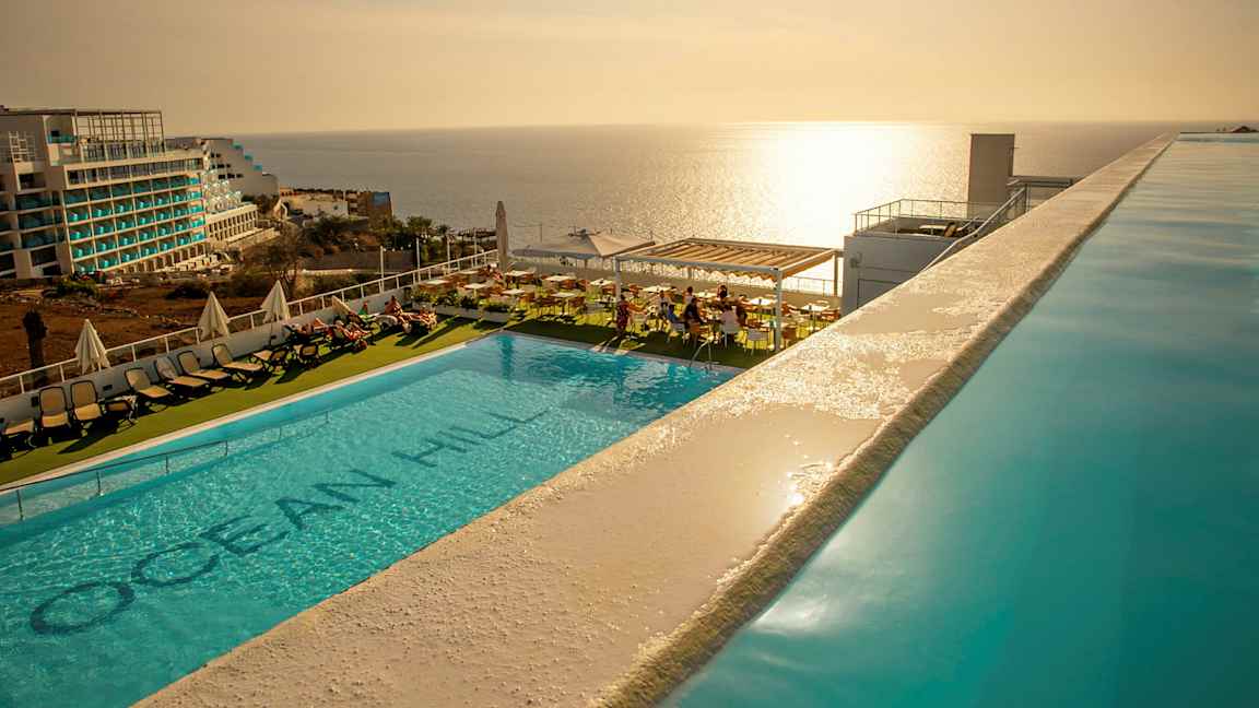 Udendørs swimmingpool med liggestole og udsigt over havet ved solnedgang. (AI-genereret tekst)