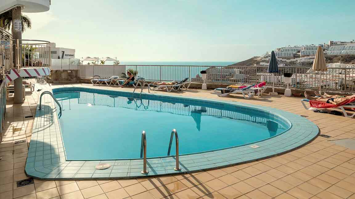 En udendørs swimmingpool med liggestole og parasoller omkring, med udsigt over havet og omkringliggende bygninger i baggrunden. (AI-genereret tekst)