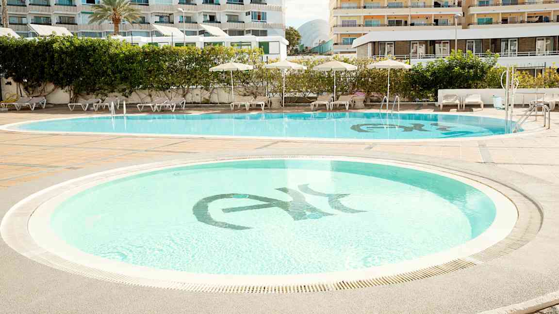 To swimmingpools omgivet af liggestole og parasoller, med hotelbygninger i baggrunden. (AI-genereret tekst)