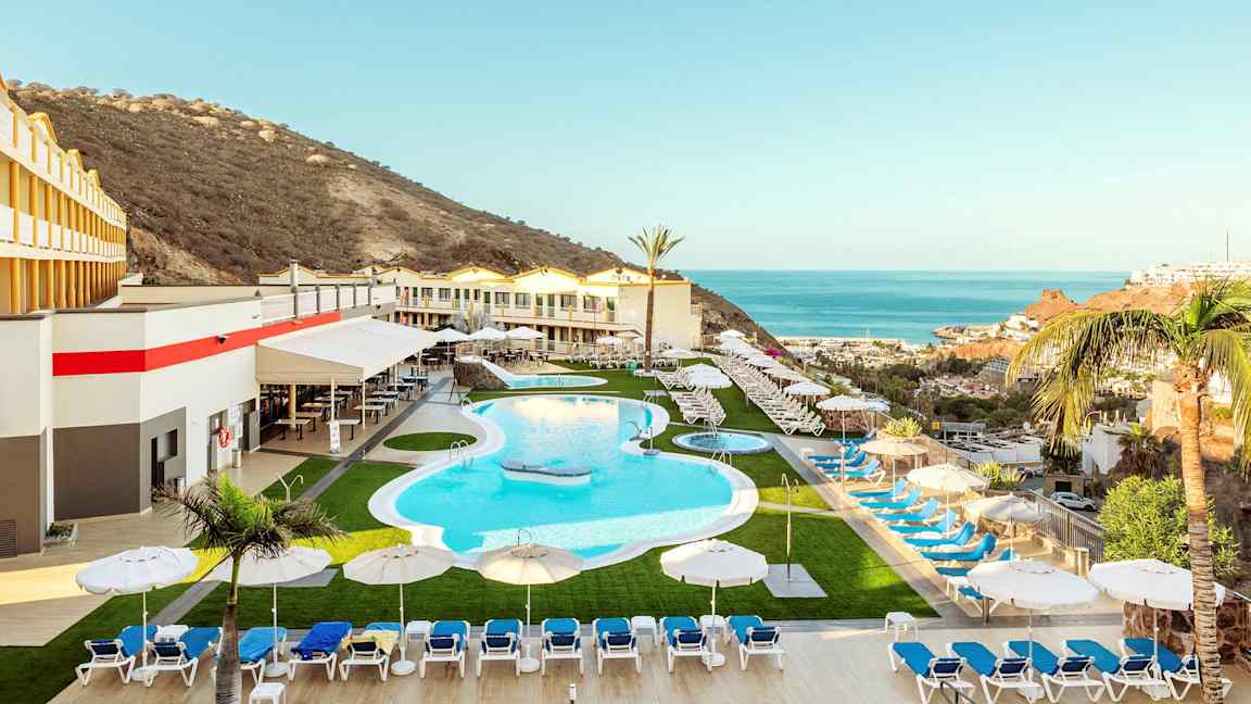 Et feriehotel med en pool, liggestole og parasoller, omgivet af et bjerglandskab og udsigt over havet. (AI-genereret tekst)