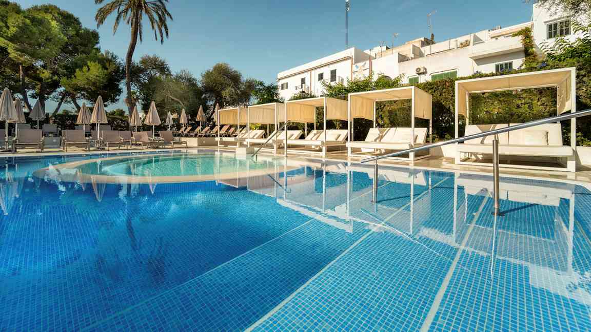 Billede af en pool med turkis vand samt hvide solsenge under parasoller og cabanas. Der er omkringliggende grønt og en skygge af bygninger i baggrunden. (AI-genereret tekst)
