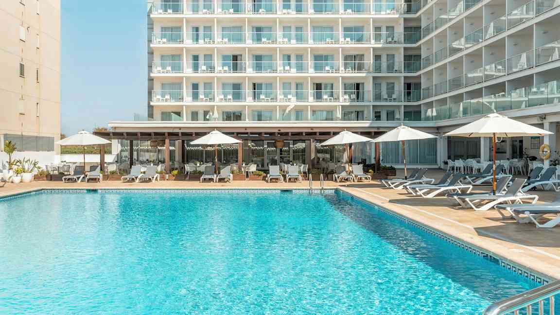 En bygning med flere altaner, foran en pool med liggestole og parasoller. (AI-genereret tekst)