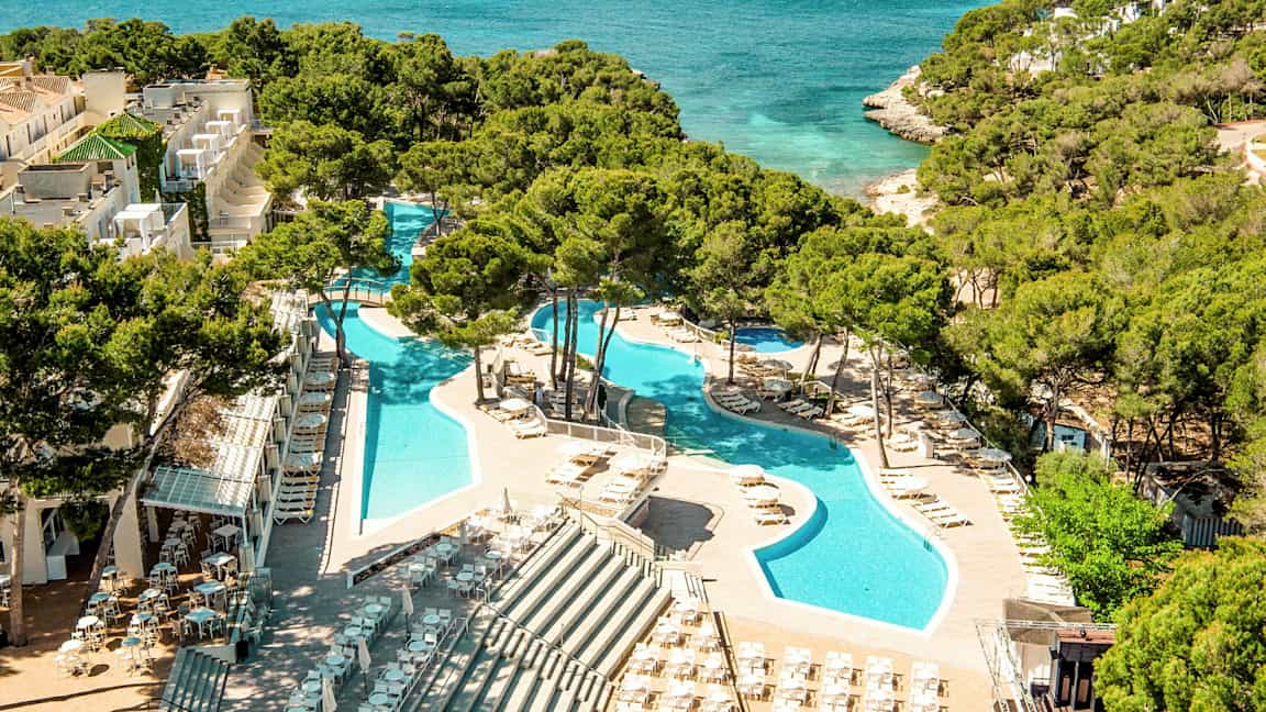 Udsigt over en ferieanlæg med swimmingpools omgivet af liggestole og parasoller, beliggende ved siden af et turkisblåt hav med frodig vegetation i baggrunden. (AI-genereret tekst)