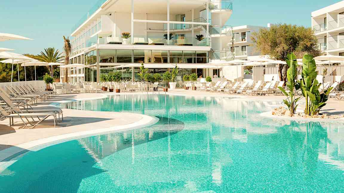 Et moderne hotel med glasaltaner ligger ved en klarblå pool omgivet af liggestole og grønne planter. (AI-genereret tekst)