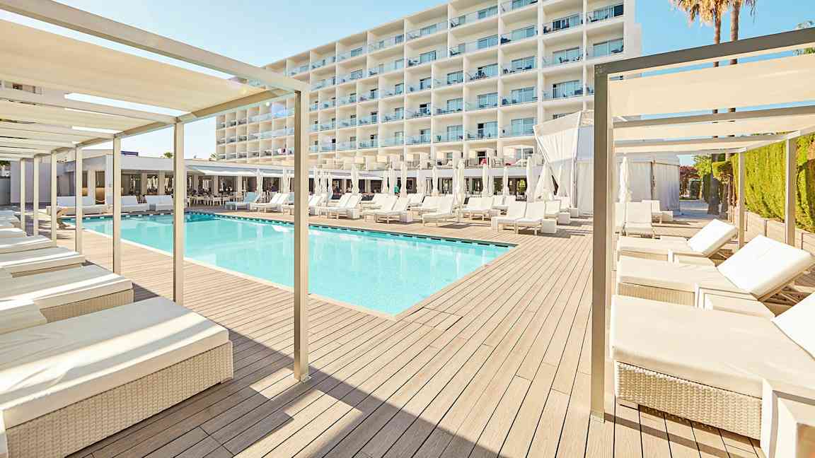 Et moderne hotelldesign med en pool omgivet af liggestole og parasoller, med en stor hvid bygning i baggrunden. (AI-genereret tekst)