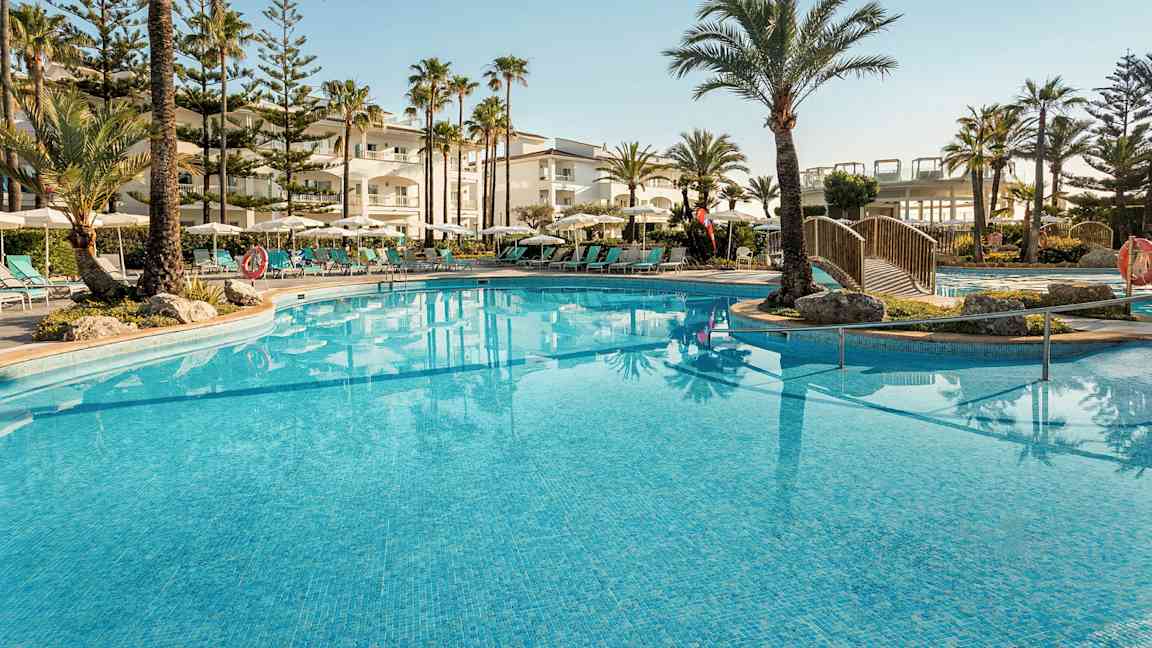 En udendørs pool omgivet af liggestole, parasoller og palmer ved et hotel. (AI-genereret tekst)