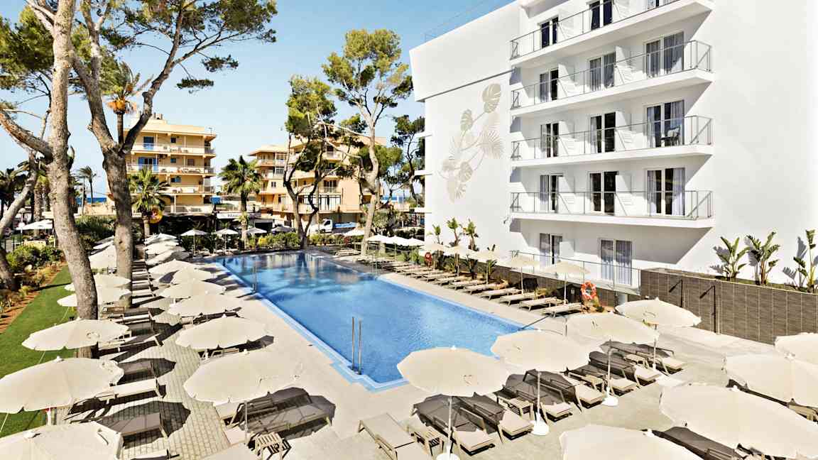 Billede af et hotelområde med en stor pool omgivet af solsenge og parasoller. En hvid bygning med flere altaner samt træer i baggrunden. (AI-genereret tekst)