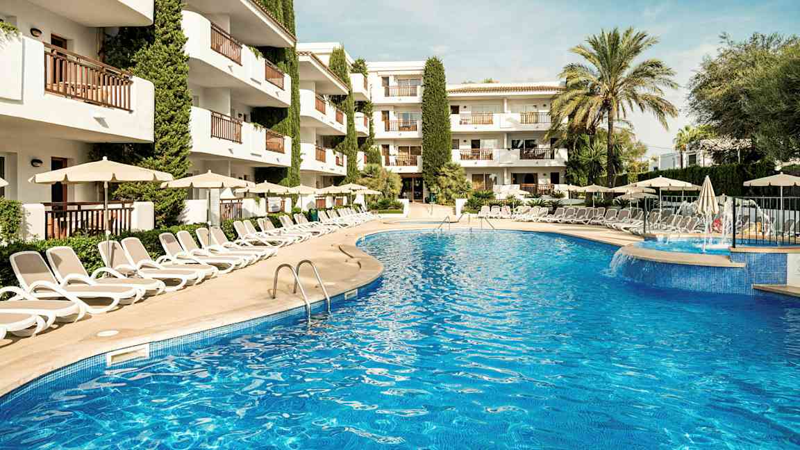 Billede af en udendørs pool omgivet af liggestole og parasoller, med et hotel i baggrunden. (AI-genereret tekst)